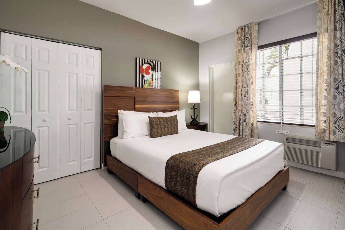 A neatly arranged bedroom with a wooden bed, white bedding, a bedside table with a lamp, and a window with patterned curtains.