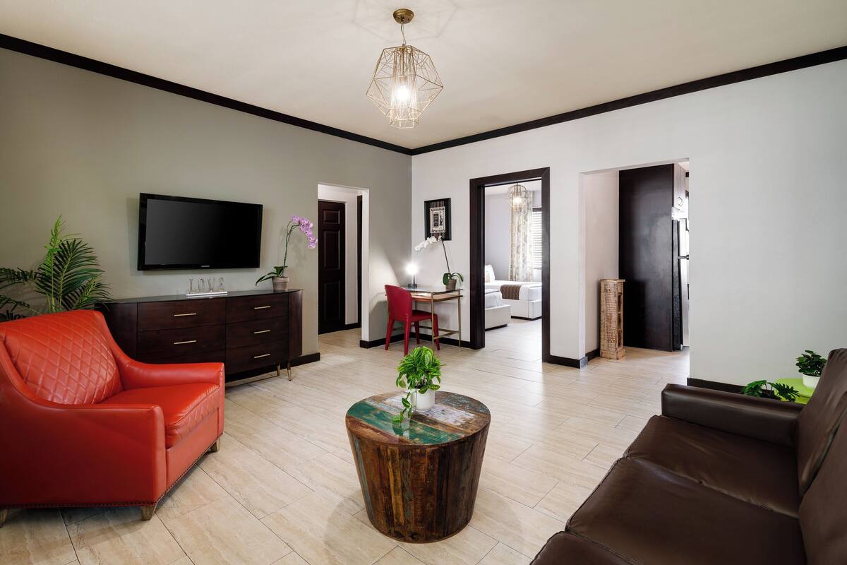 A modern living room with a flat-screen TV, a red leather sofa, a brown leather sofa, a wooden coffee table with a plant, and a dining area with a red chair.