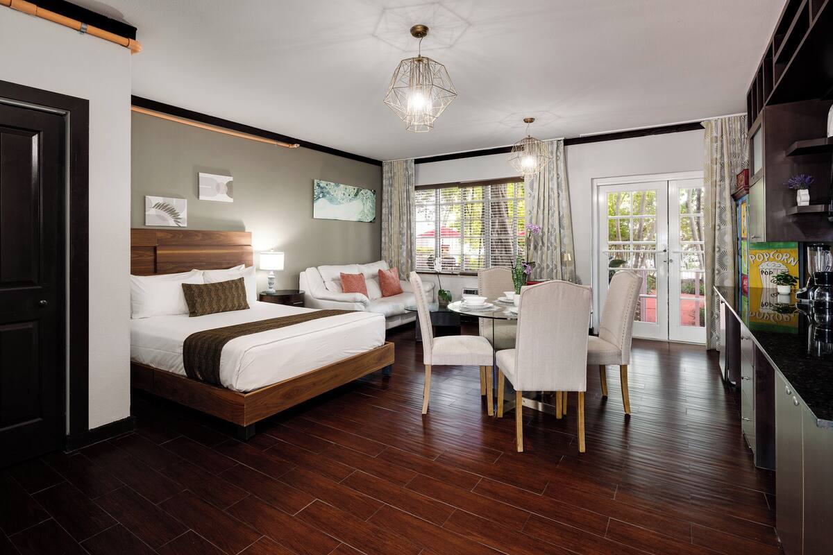 A modern hotel room with a wooden bed, a dining table with chairs, and a kitchen area.