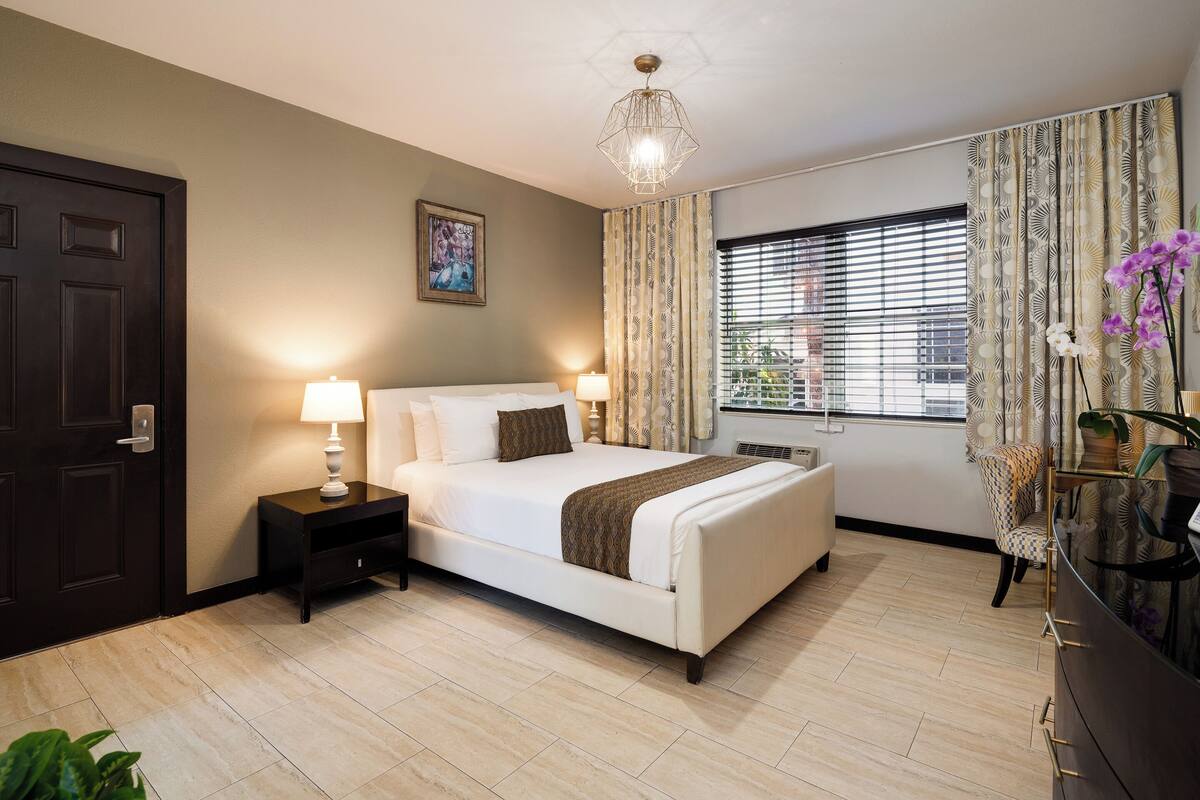 A bedroom with a bed, bedside lamps, a window with blinds, a door, and a plant.