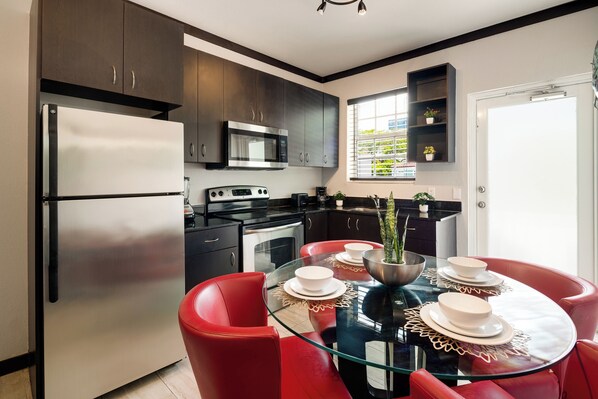 A modern kitchen with dark cabinets, stainless steel appliances, and a glass dining table with red chairs.