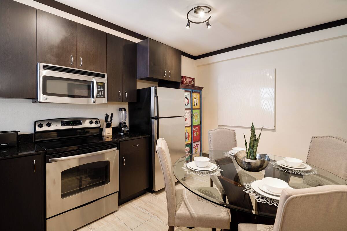 A modern kitchen with dark cabinets, stainless steel appliances, and a glass dining table set for four.