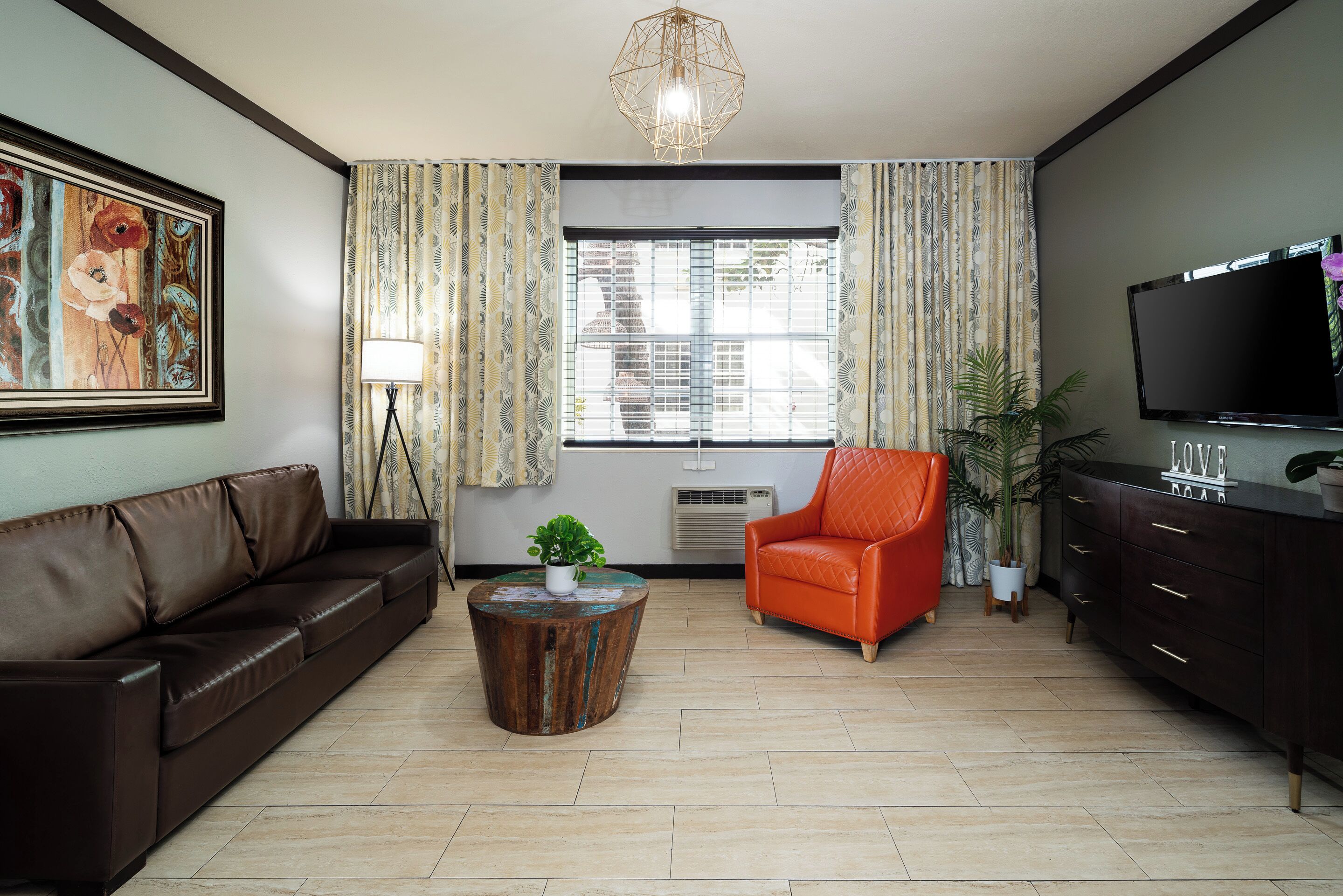 A living room with a brown leather sofa, an orange armchair, a wooden coffee table, a TV, and a large window with curtains.