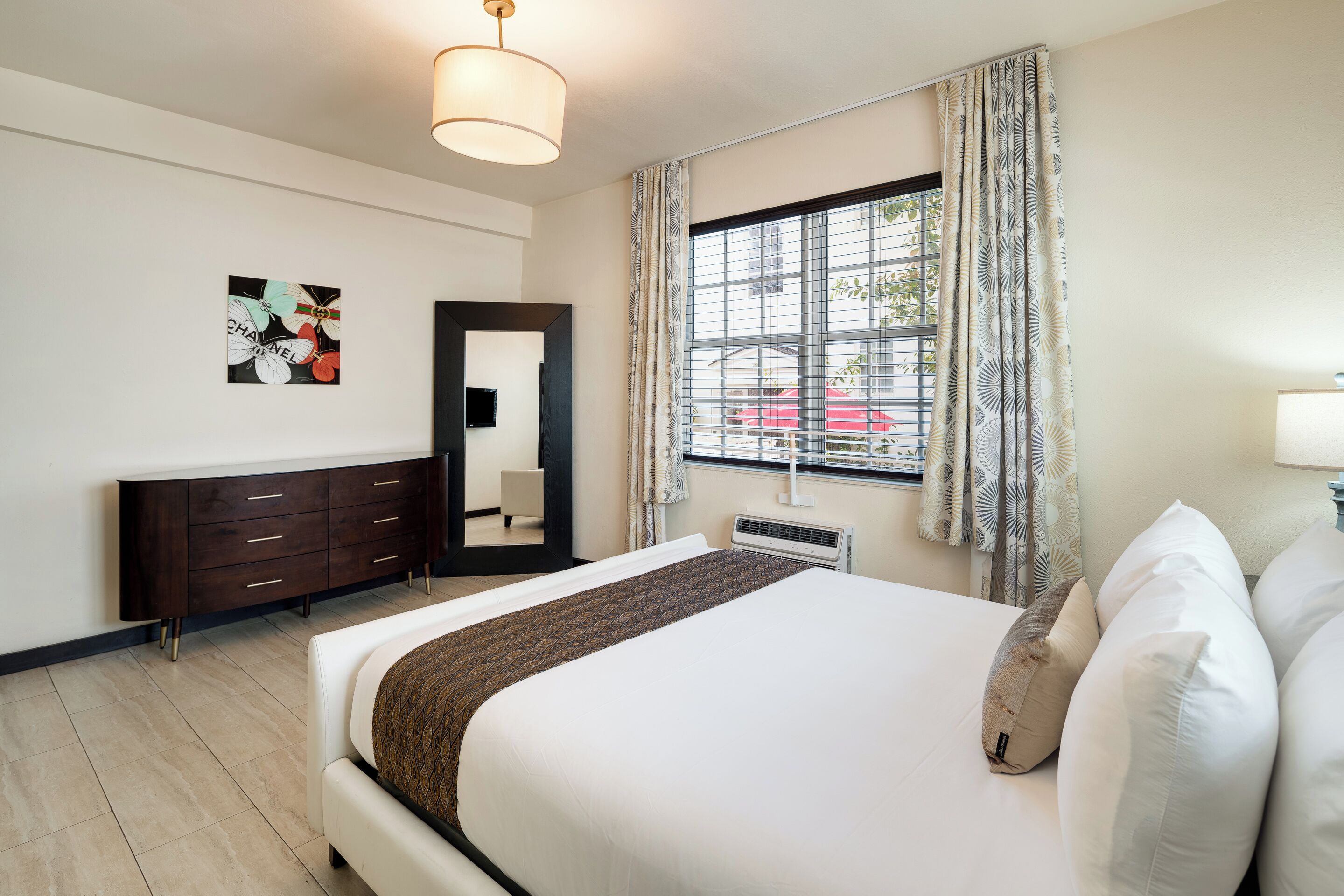 Ein Hotelzimmer mit einem großen Bett, einem Schminktisch, einem Spiegel, einem Fenster mit Vorhängen und einem an der Wand hängenden Kunstwerk.
