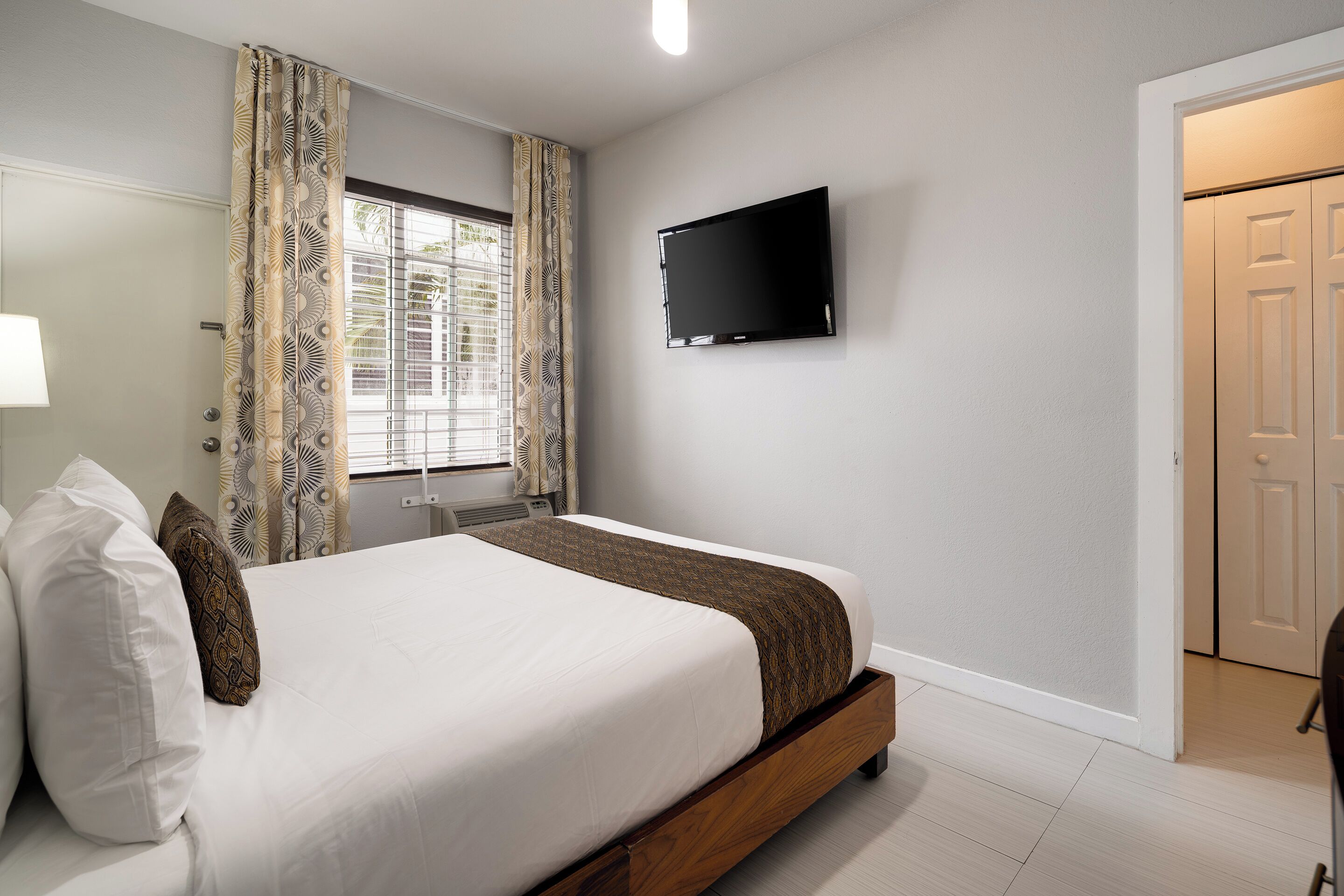 Un dormitorio con una cama, un televisor montado en la pared y una ventana con cortinas.