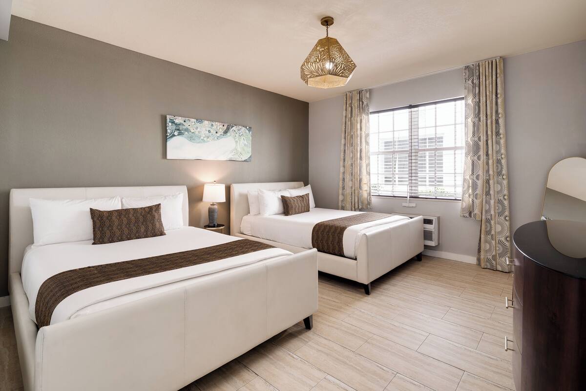 A hotel room with two beds, a large window with curtains, a ceiling light, and a wall-mounted artwork.