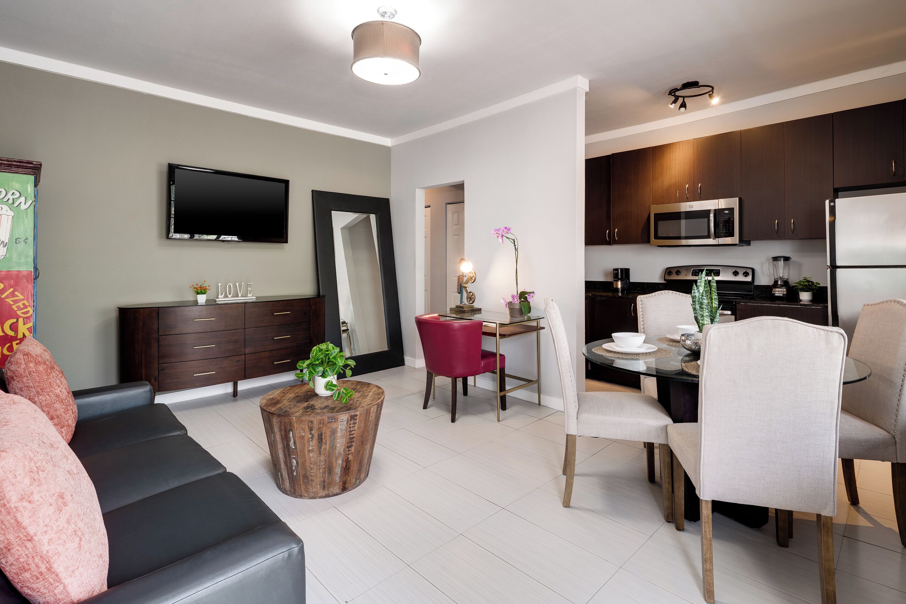 A modern living room with a sofa, a dining table with chairs, a kitchen area with appliances, and a flat-screen TV mounted on the wall.