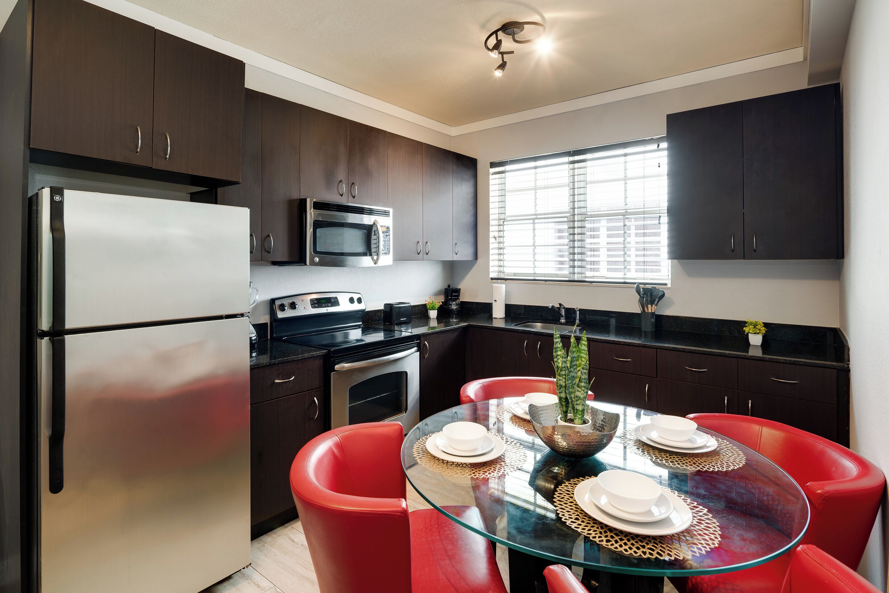 A modern kitchen with dark wood cabinets, stainless steel appliances, and a glass dining table with red chairs.
