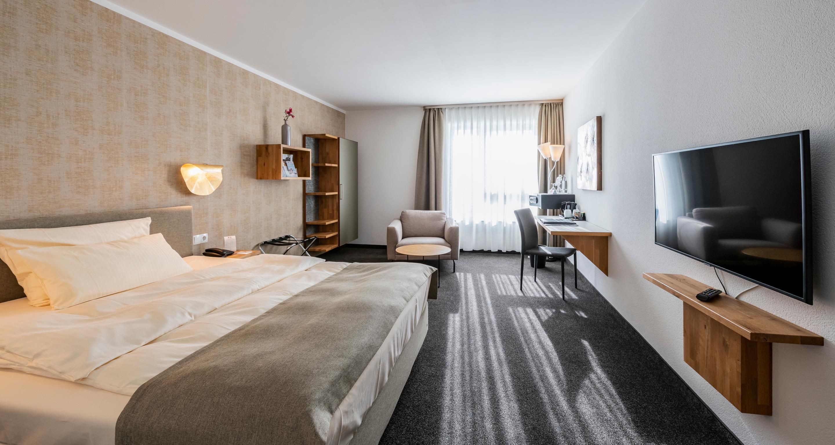 A hotel room with a bed, a desk, a chair, a TV, and a bookshelf.