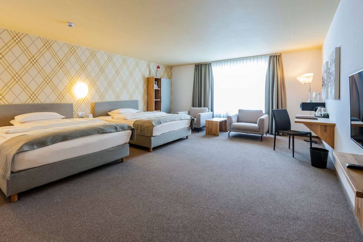 Ein Hotelzimmer mit zwei Betten, einem Schreibtisch, einem Stuhl, einer Lampe und einem großen Fenster.