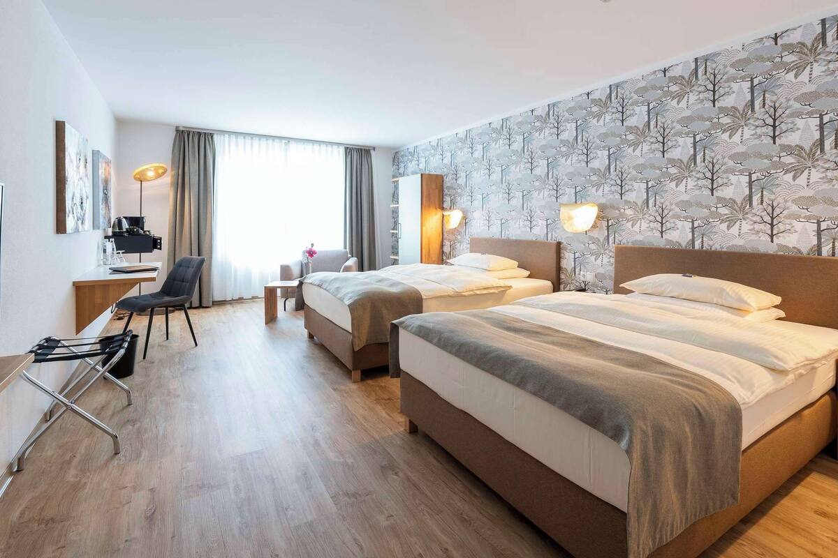 Ein Hotelzimmer mit zwei Betten, einem Schreibtisch, einem Sessel, einem Fernseher und einem großen Fenster mit Vorhängen.