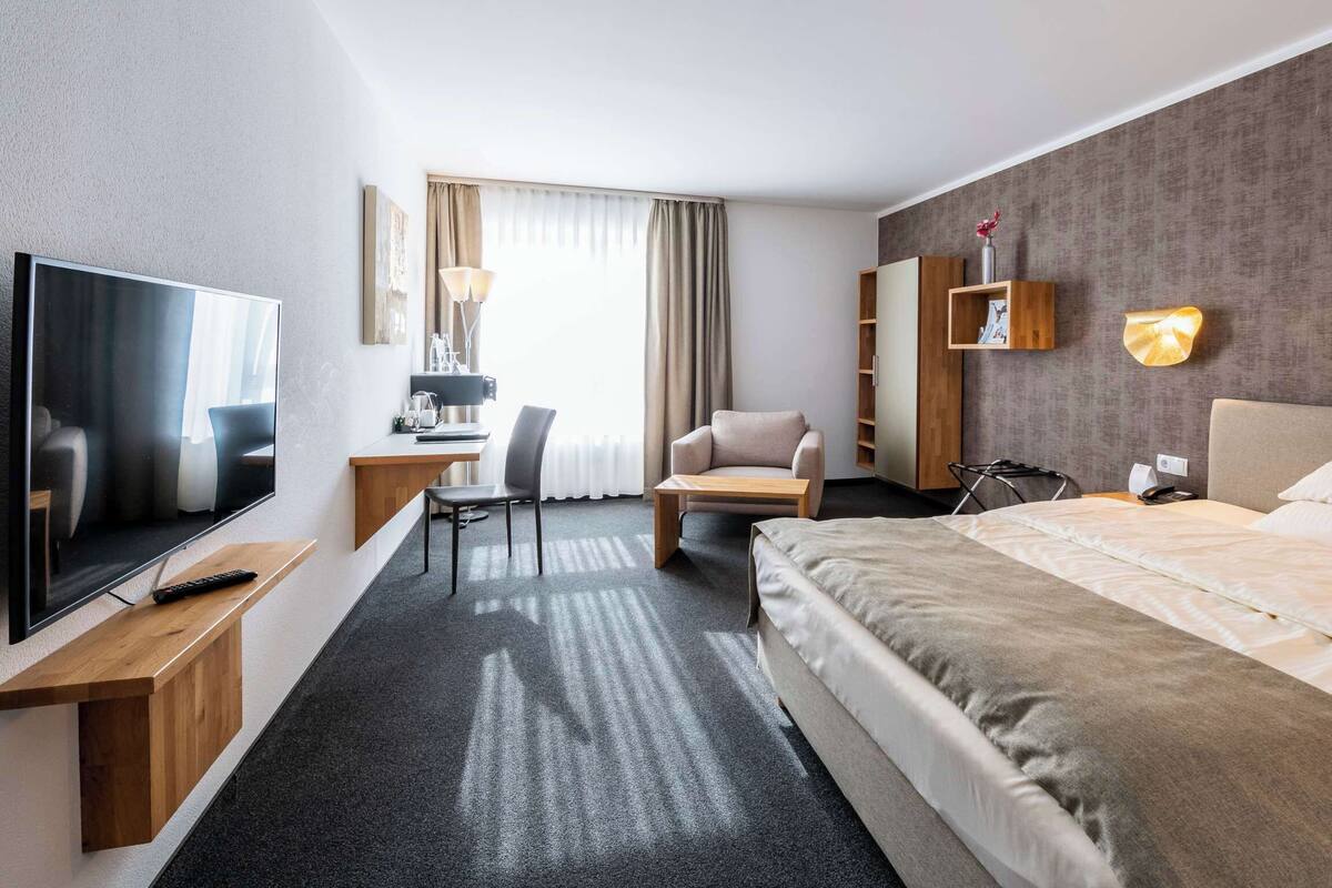 Ein Hotelzimmer mit einem großen Bett, einem Schreibtisch mit Stuhl, einem Fernseher und einem an der Wand befestigten Regal mit einer Vase.