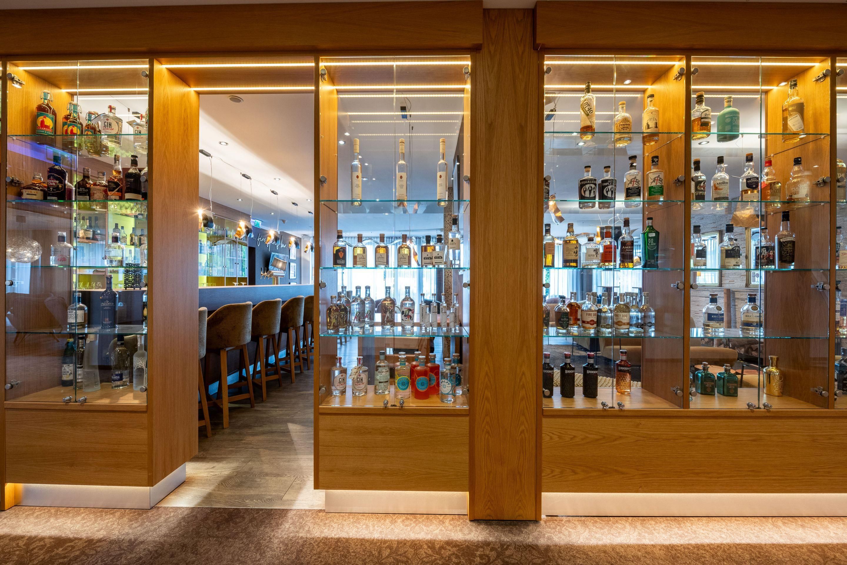 A bar area with glass shelving displaying various liquor bottles, wooden furniture, and a carpeted floor.