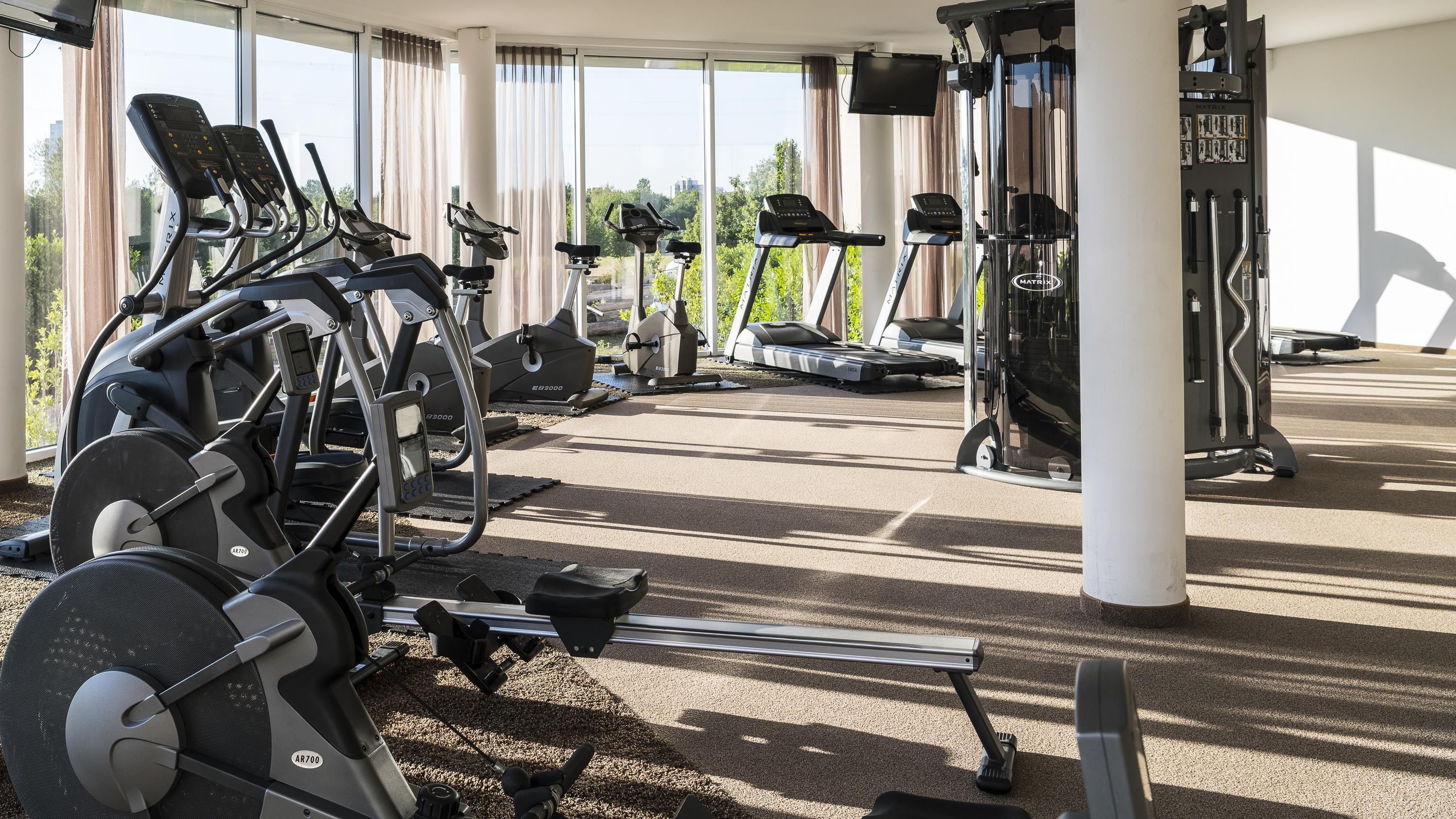 Une salle de sport équipée de divers appareils d’exercice, notamment des tapis de course et un rameur, située dans un espace abrité doté de grandes fenêtres.