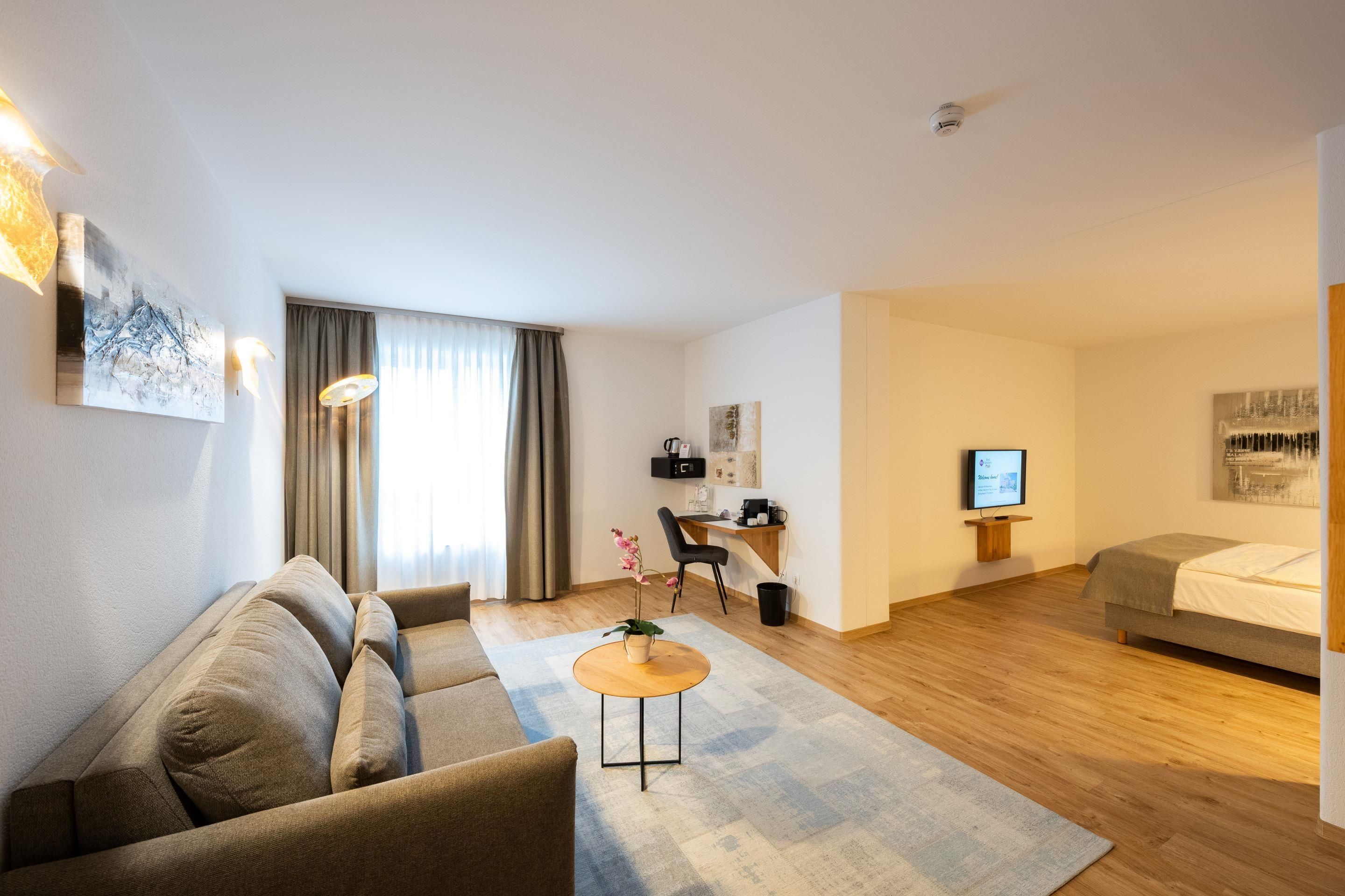 Quarto de hotel moderno com sofá, mesa redonda pequena, escrivaninha com cadeira, cama e televisão.