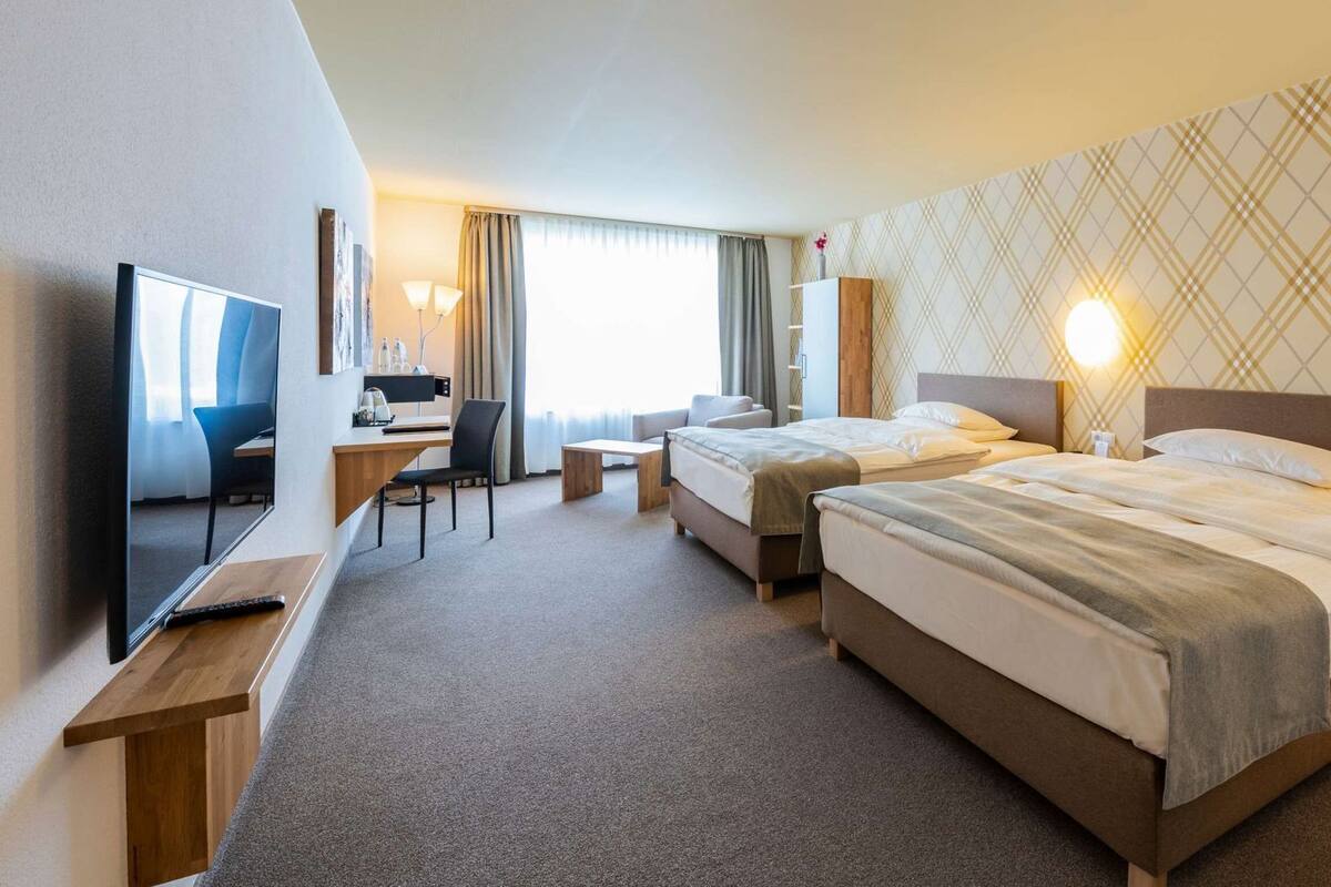 Ein Hotelzimmer mit zwei Betten, einem Schreibtisch, einem Sessel, einem Fernseher und einem großen Fenster.