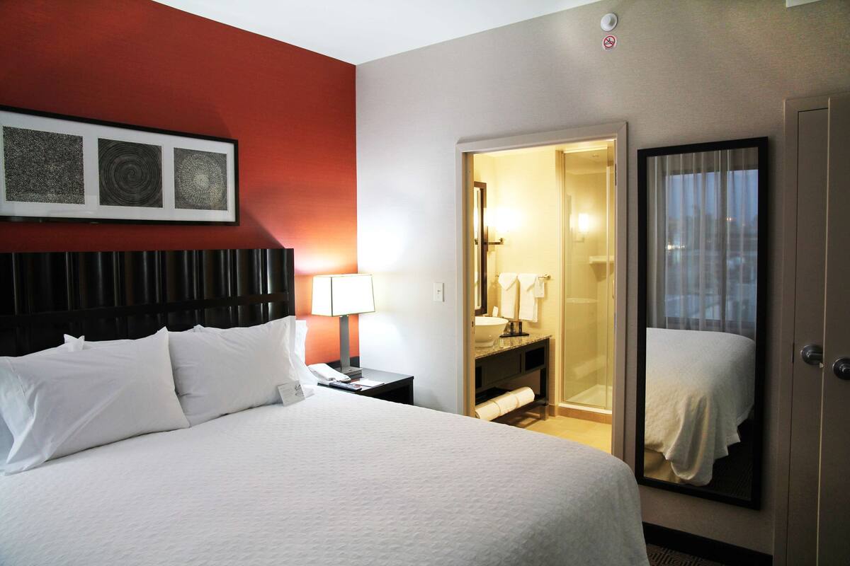 A hotel room with a bed, a nightstand, a lamp, a bathroom visible through an open door, and a framed picture on the wall.