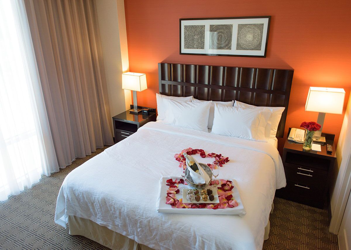Habitación de hotel con una cama grande, dos mesitas de noche con lámparas, una obra de arte enmarcada sobre la cama y una bandeja con una botella y bombones en la cama.