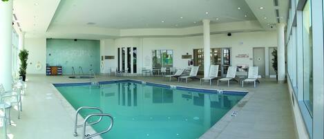Una piscina interior con agua cristalina, rodeada de sillas de descanso y un suelo de baldosas.