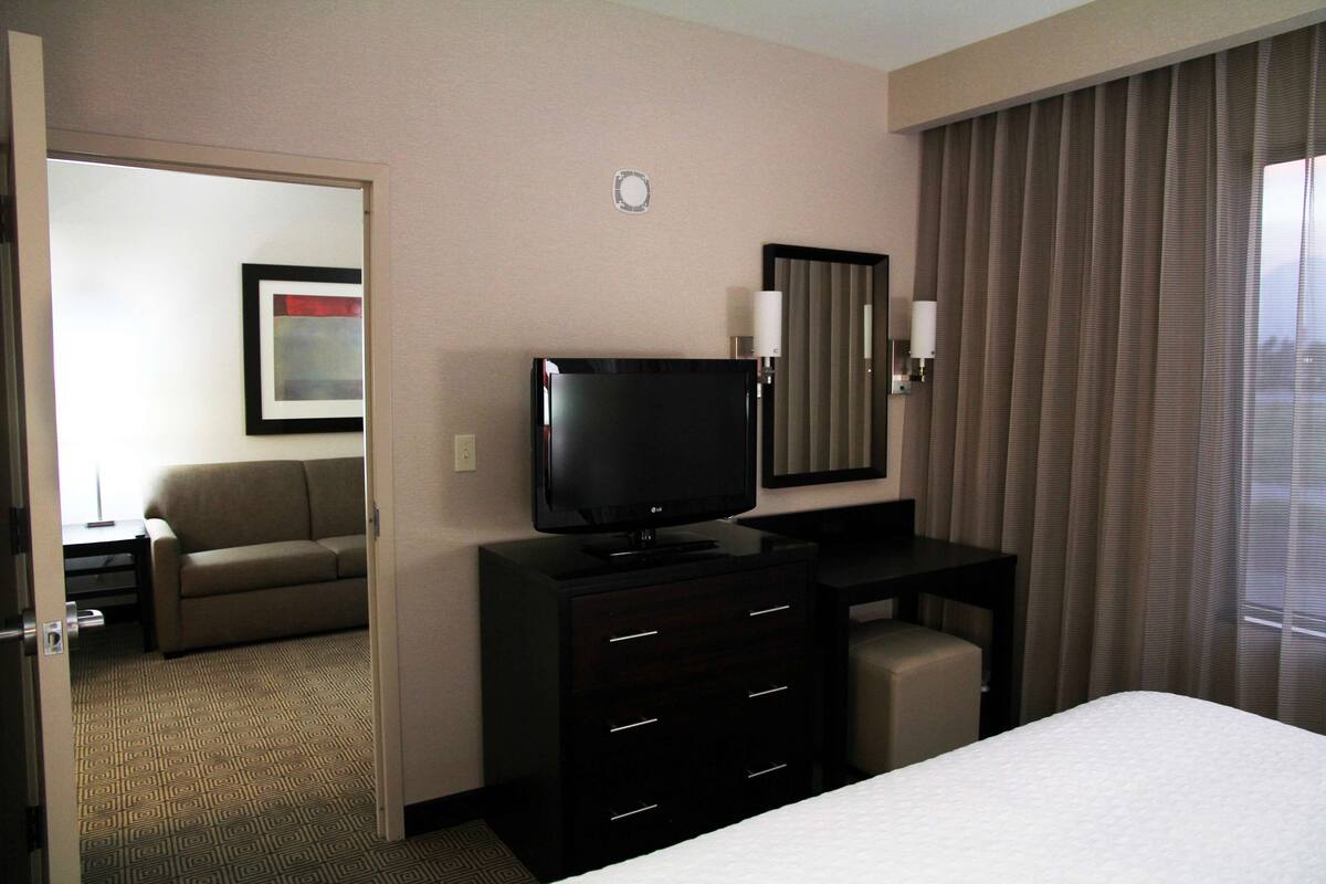 A hotel room with a bed, a television, a desk, a chair, a dresser, a mirror, a window with curtains, and a door leading to another room.