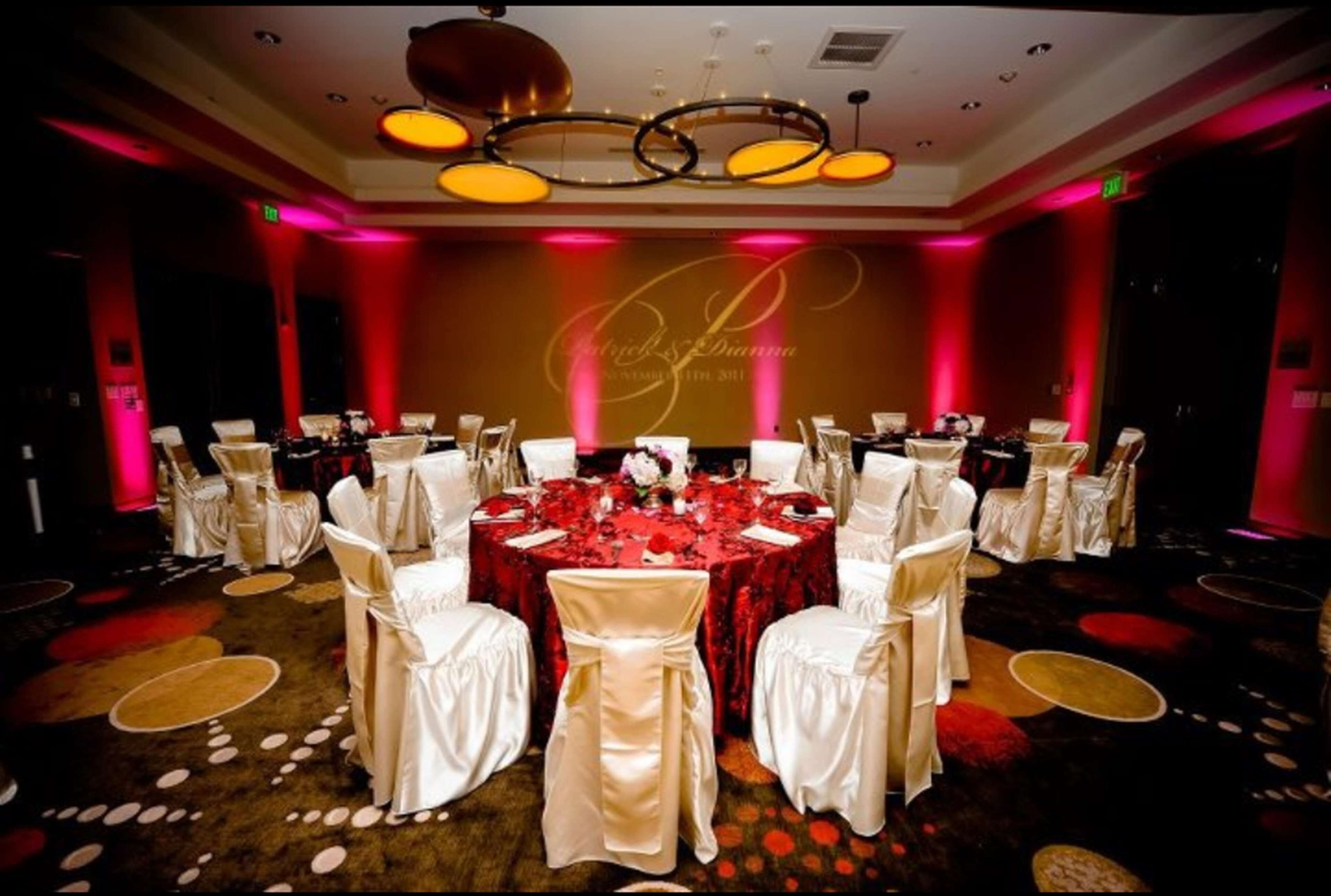 Un salón de banquetes con mesas redondas dispuestas para un evento formal, que incluye una alfombra roja y sillas blancas.