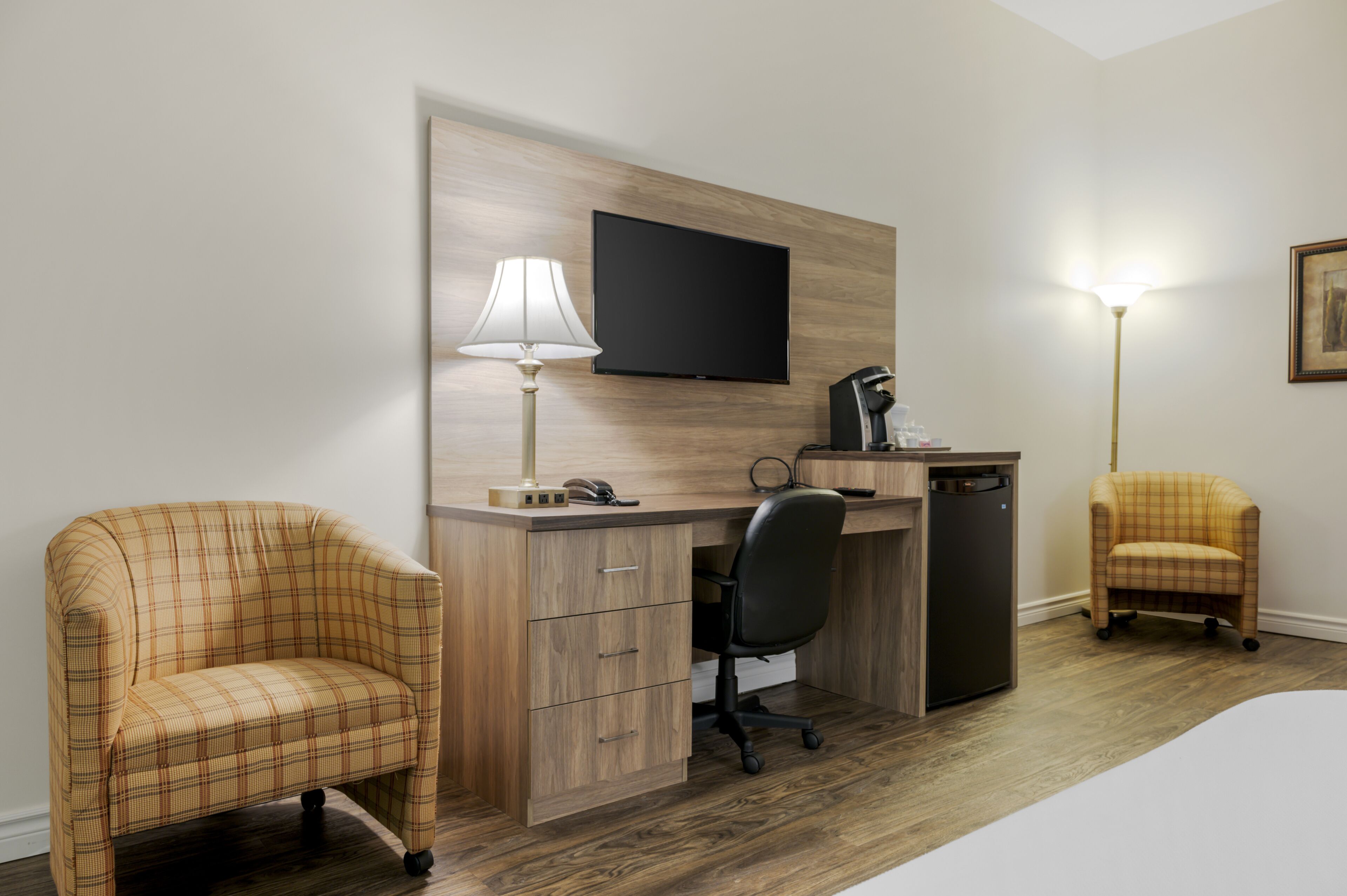 A hotel room with a wooden desk, two plaid armchairs, a flat-screen TV, a lamp, a refrigerator, and a bedside table.
