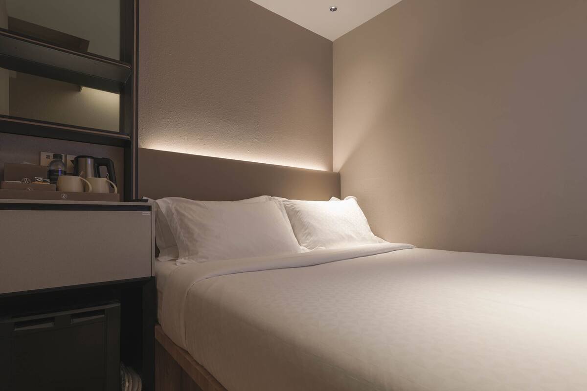 A neatly made bed in a hotel room with a headboard, a bedside table with a lamp, and a small shelf with toiletries.