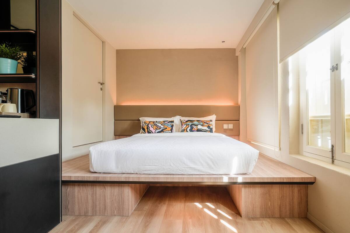 A modern bedroom with a bed centered on a wooden platform, a headboard with integrated lighting, and a large window on one side.