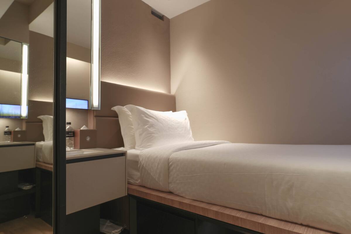 A modern hotel room with a bed, a bedside table, a mirror, and a small desk with a chair.