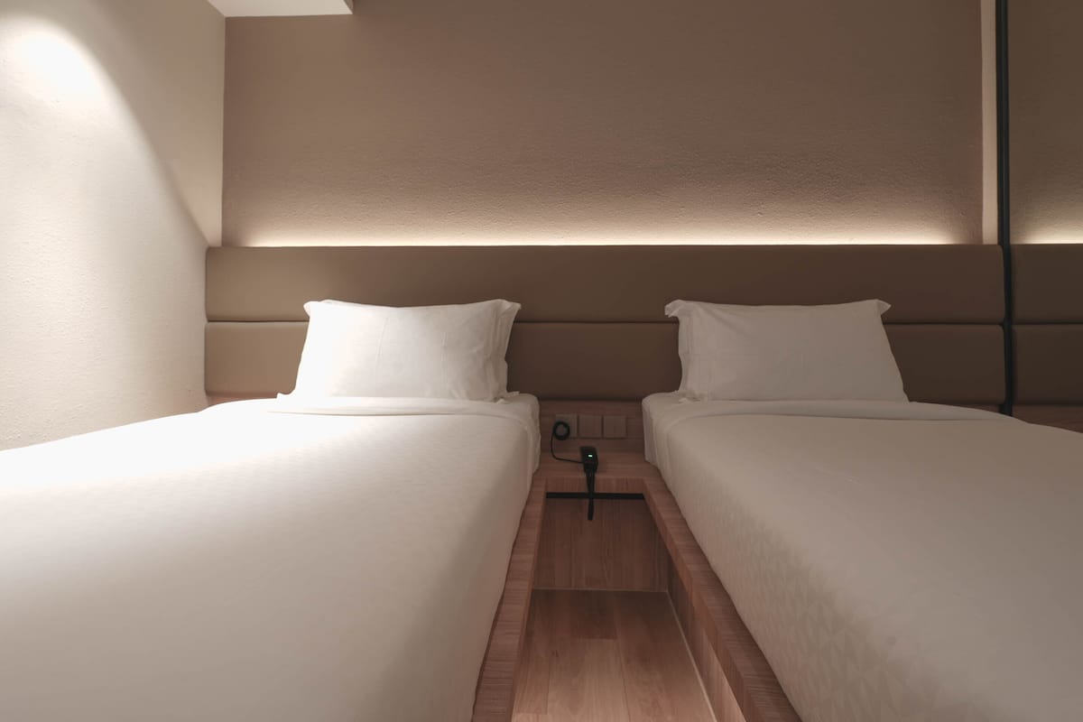 A modern hotel room with two single beds, a nightstand, and a wall-mounted lamp.