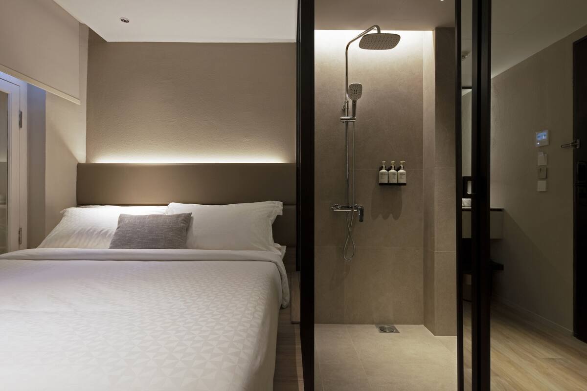 A modern hotel room with a large bed, a glass-enclosed shower, and a bedside table with toiletries.