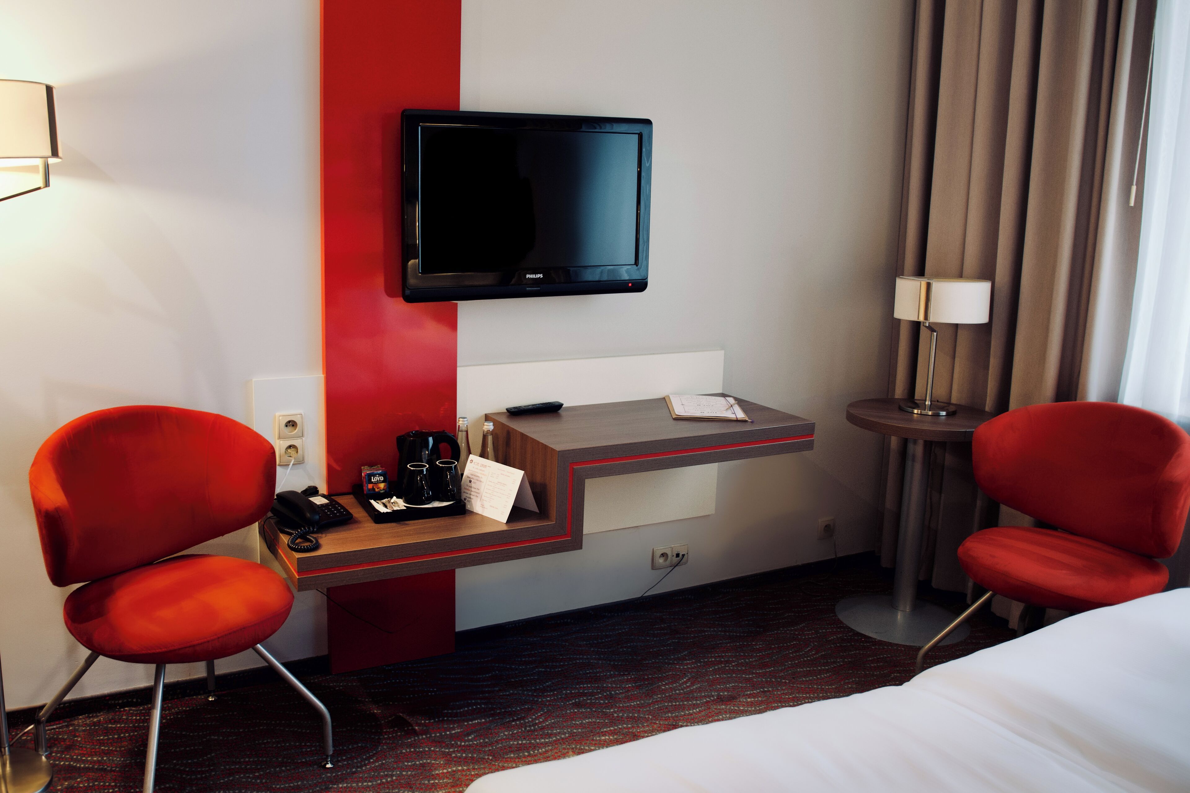 Habitación de hotel con cama, dos sillas rojas, un escritorio con televisor de pantalla plana, una lámpara y una mesita con control remoto y un bloc de notas.