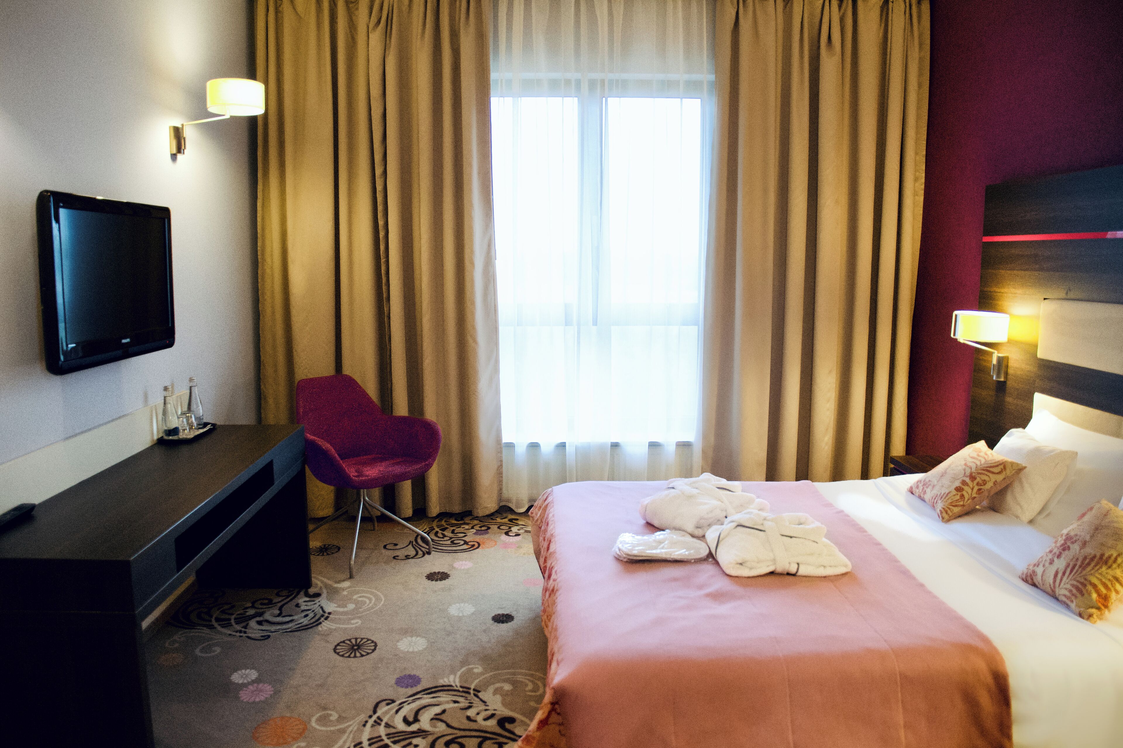 A hotel room with a bed, a desk, a chair, a TV, and a window with curtains.
