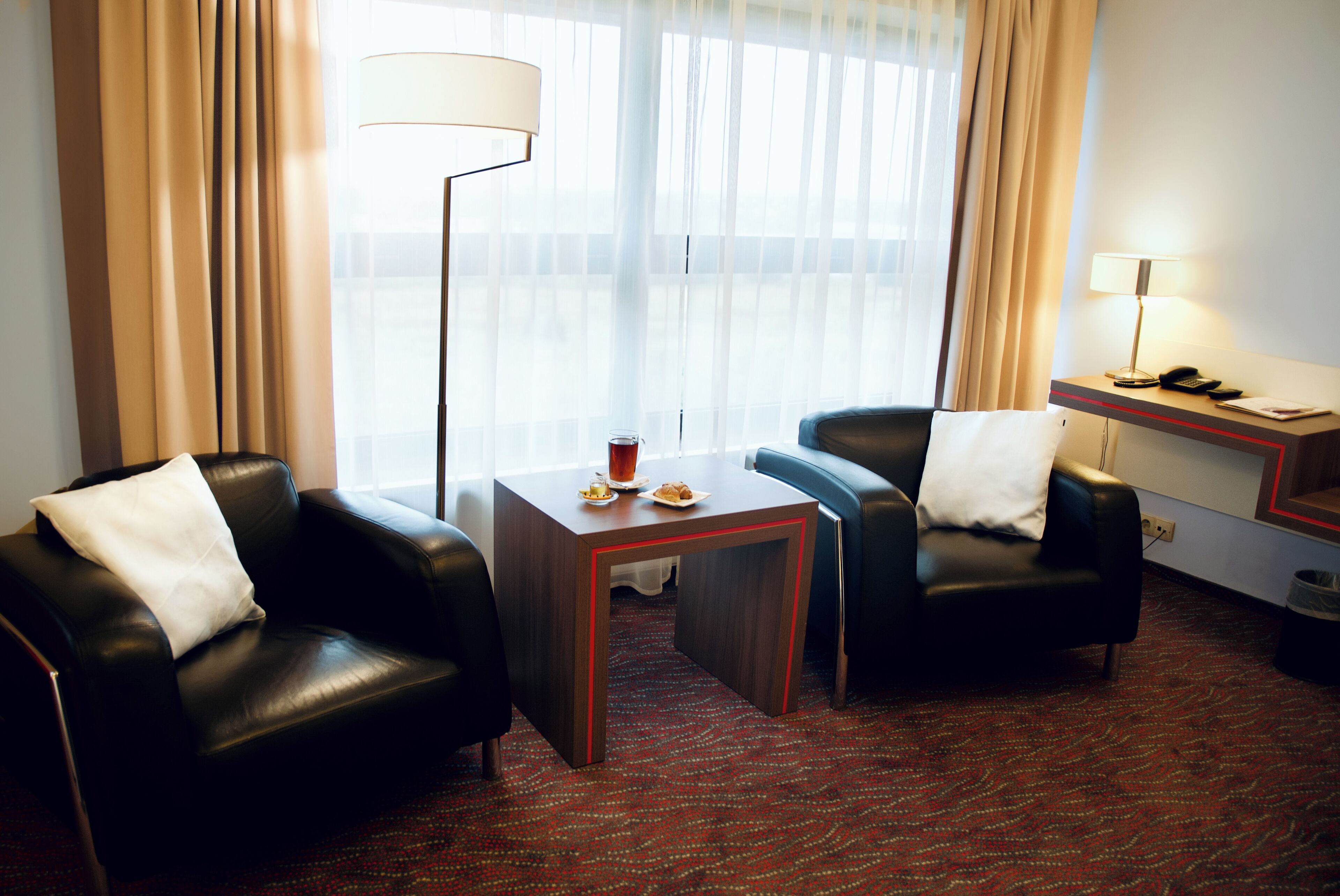 Quarto de hotel com duas poltronas de couro, uma mesinha, uma luminária e vista para o exterior através de cortinas finas.