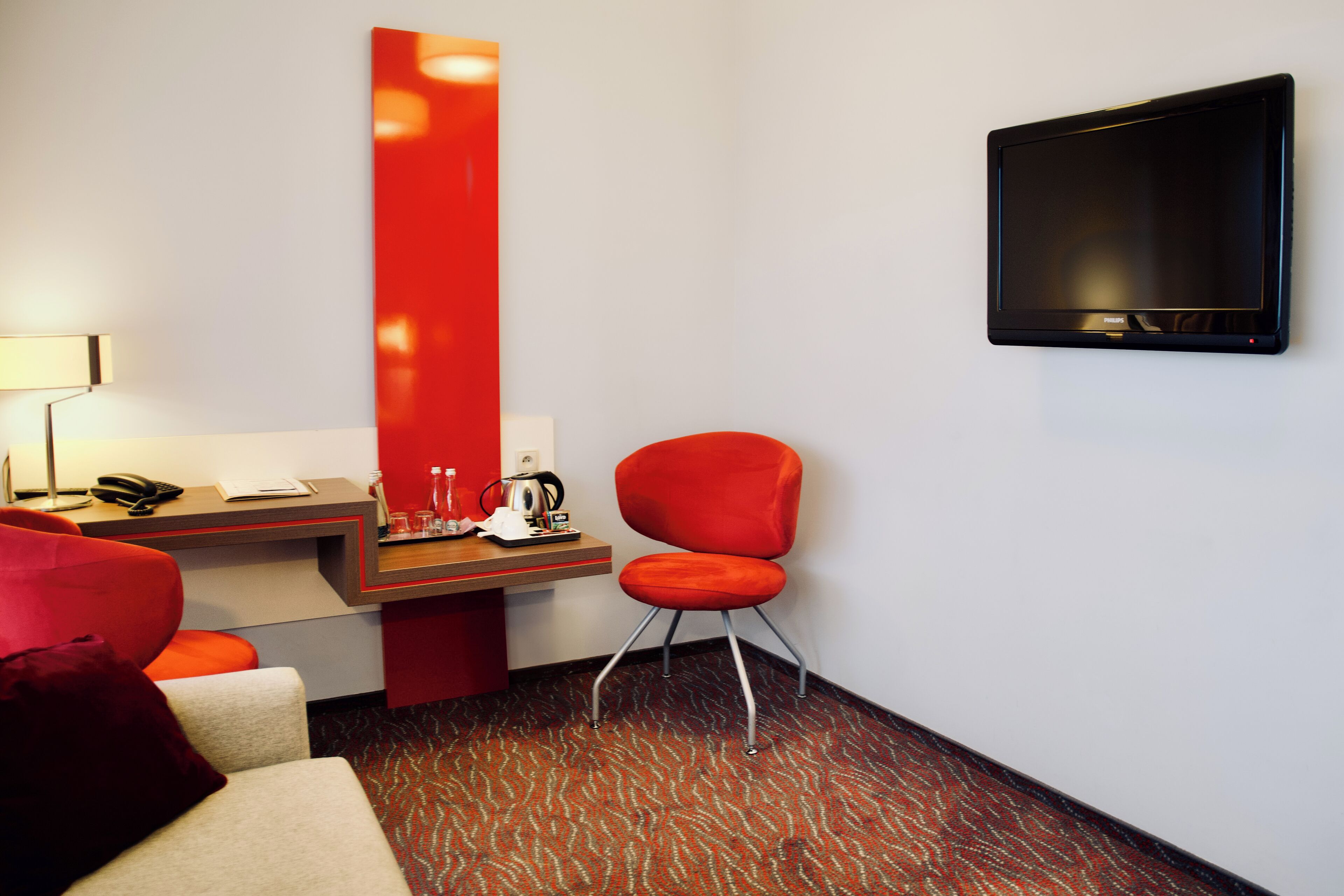 A hotel room with a desk, a red chair, a flat-screen TV mounted on the wall, and a patterned carpet.