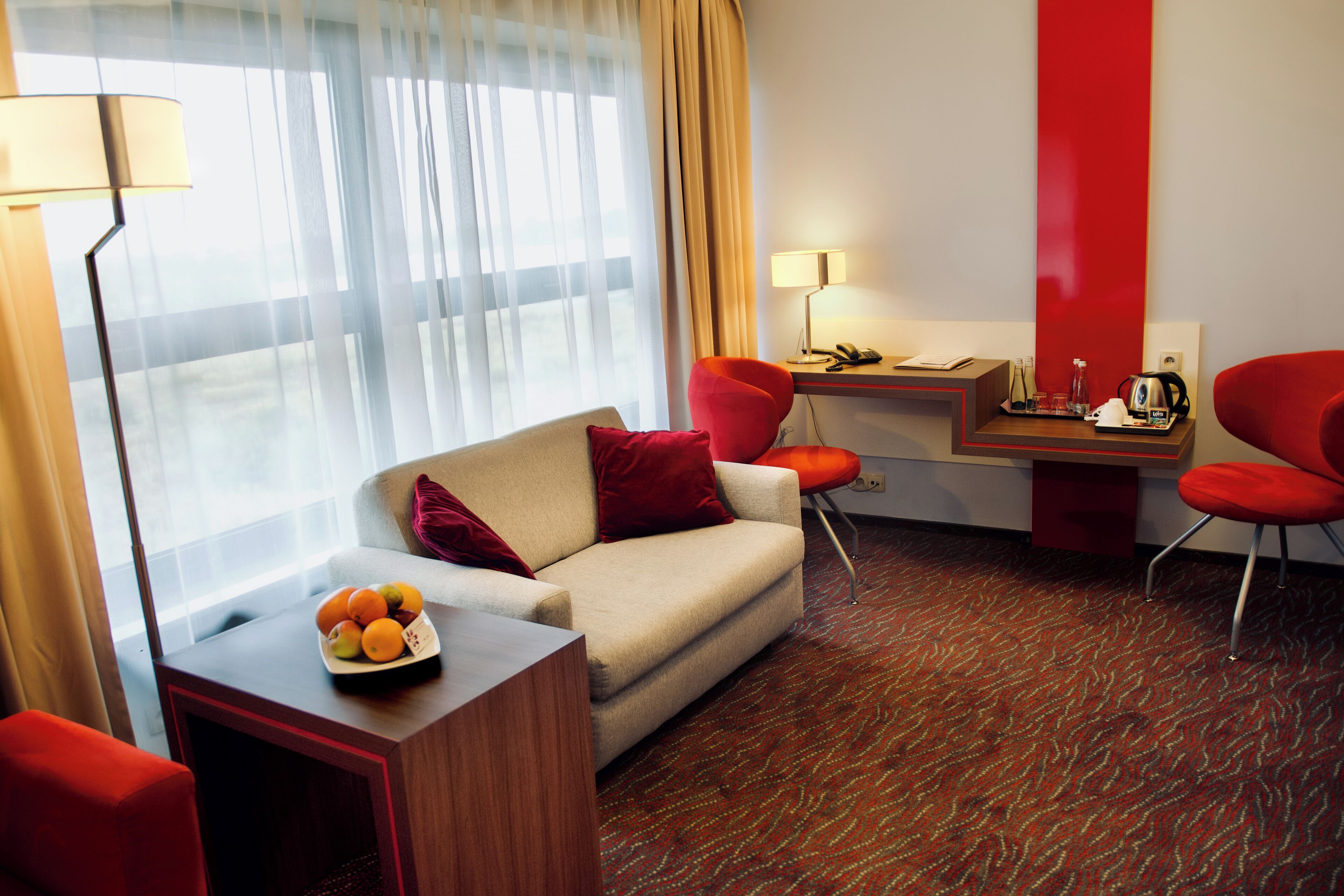 A hotel room with a sofa, a desk, and a chair. There is a fruit bowl on the desk and a lamp on the floor.