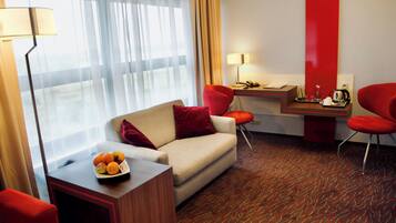 A hotel room with a sofa, a desk, and a chair. There is a fruit bowl on the desk and a lamp on the floor.