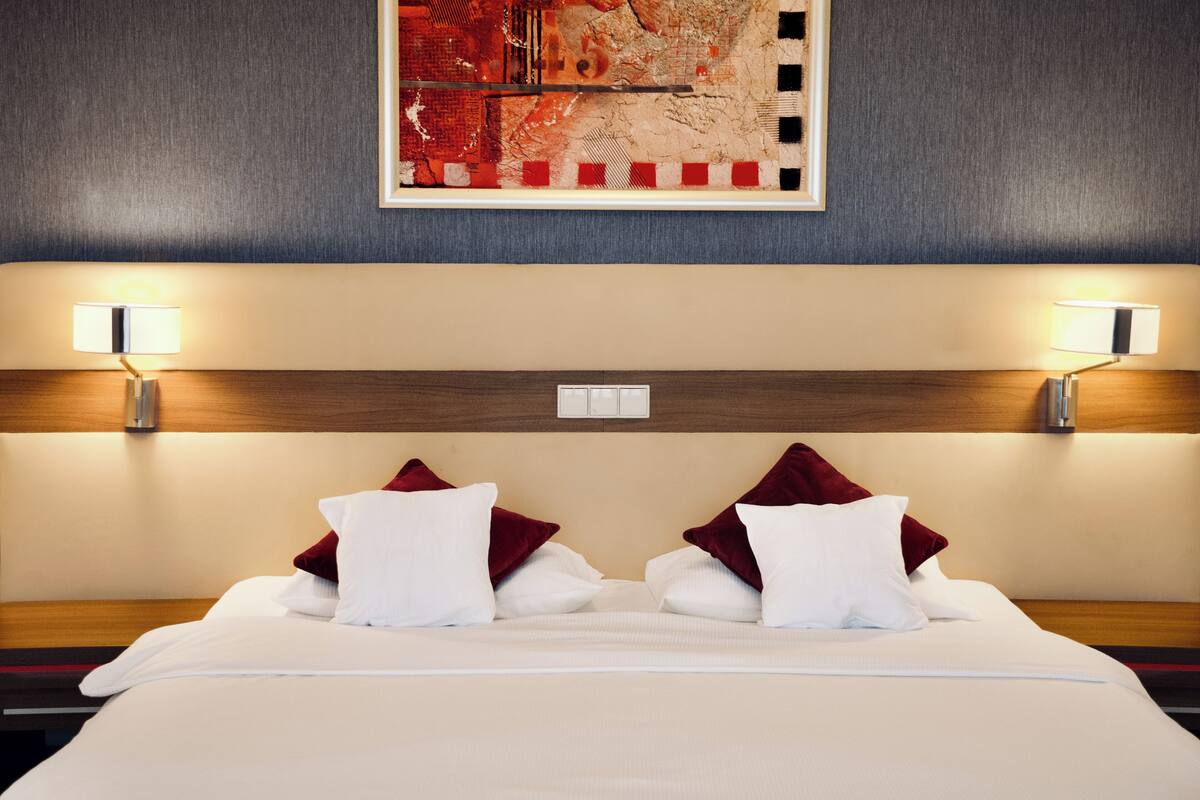 A hotel room with a neatly made bed, two wall-mounted lamps, and a framed artwork above the bed.