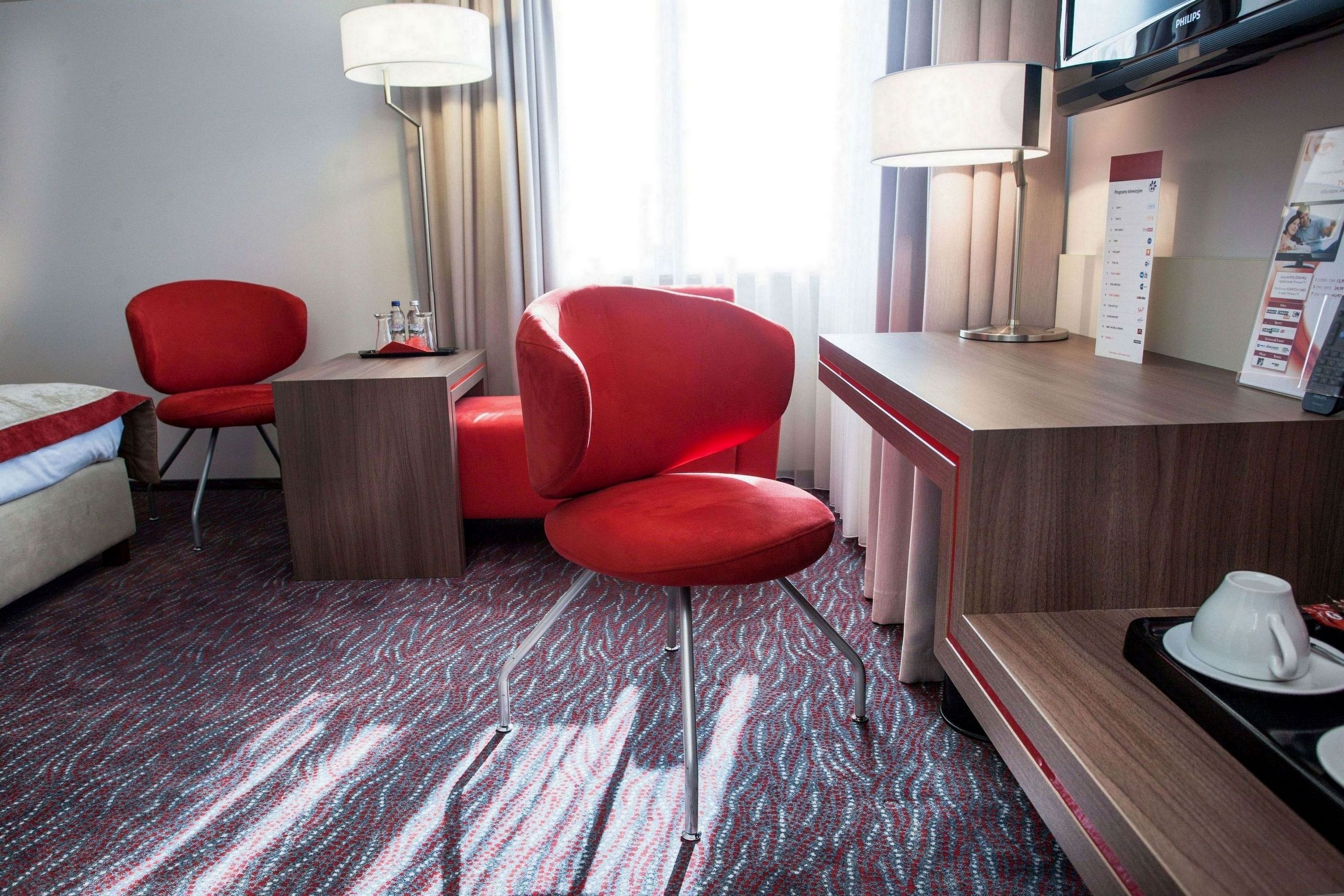 Habitación de hotel con una silla roja, un escritorio de madera, una lámpara, una cama y suelo alfombrado.