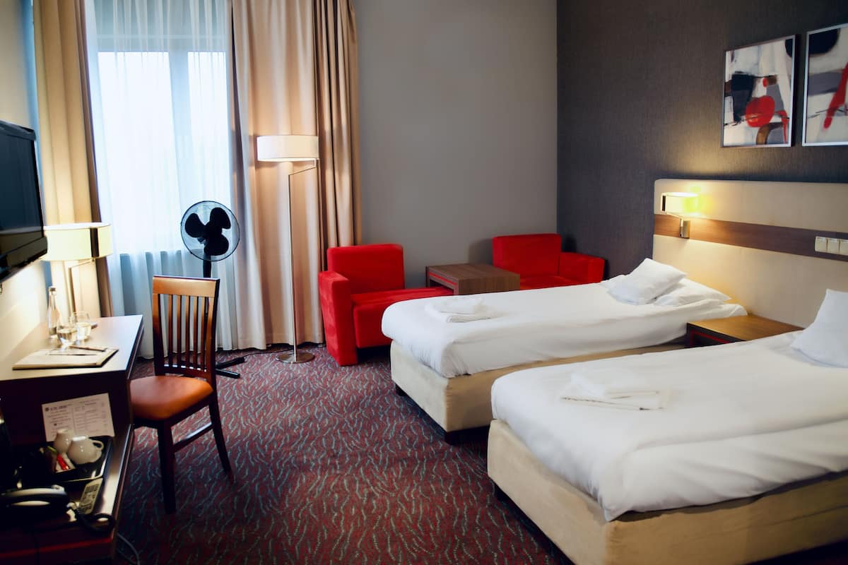 A hotel room with two beds, a desk with a lamp, a chair, a small table, and a fan.