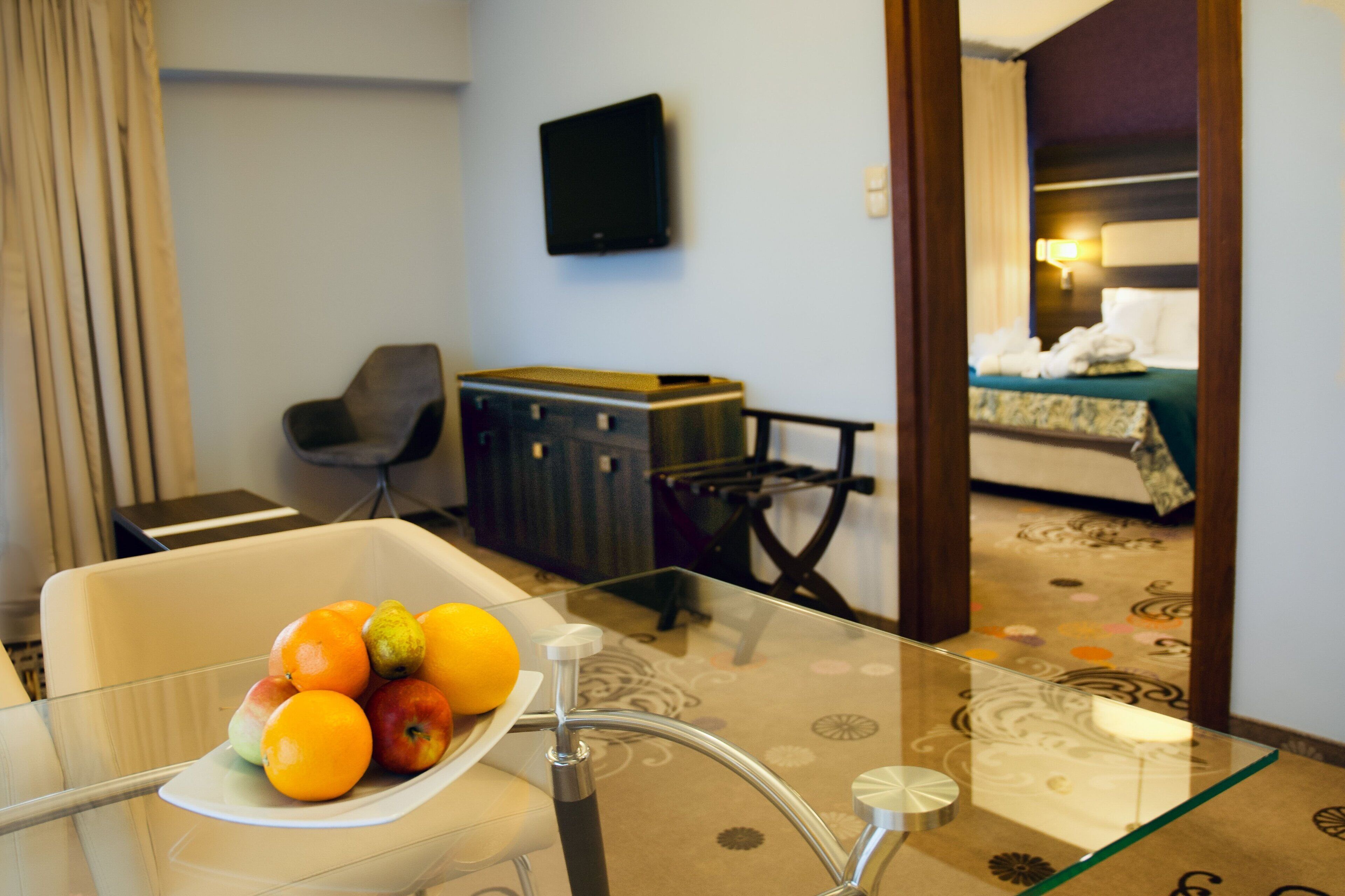 Habitación de hotel con una mesa de vidrio mostrando fruta, un televisor en la pared, una cama con cubrecama verde y una silla.