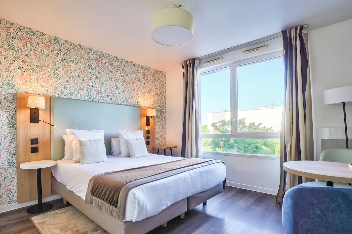Una habitación de hotel con una cama grande, dos mesitas de noche, una pequeña mesa redonda, una silla, un ventanal con cortinas y un papel tapiz estampado.