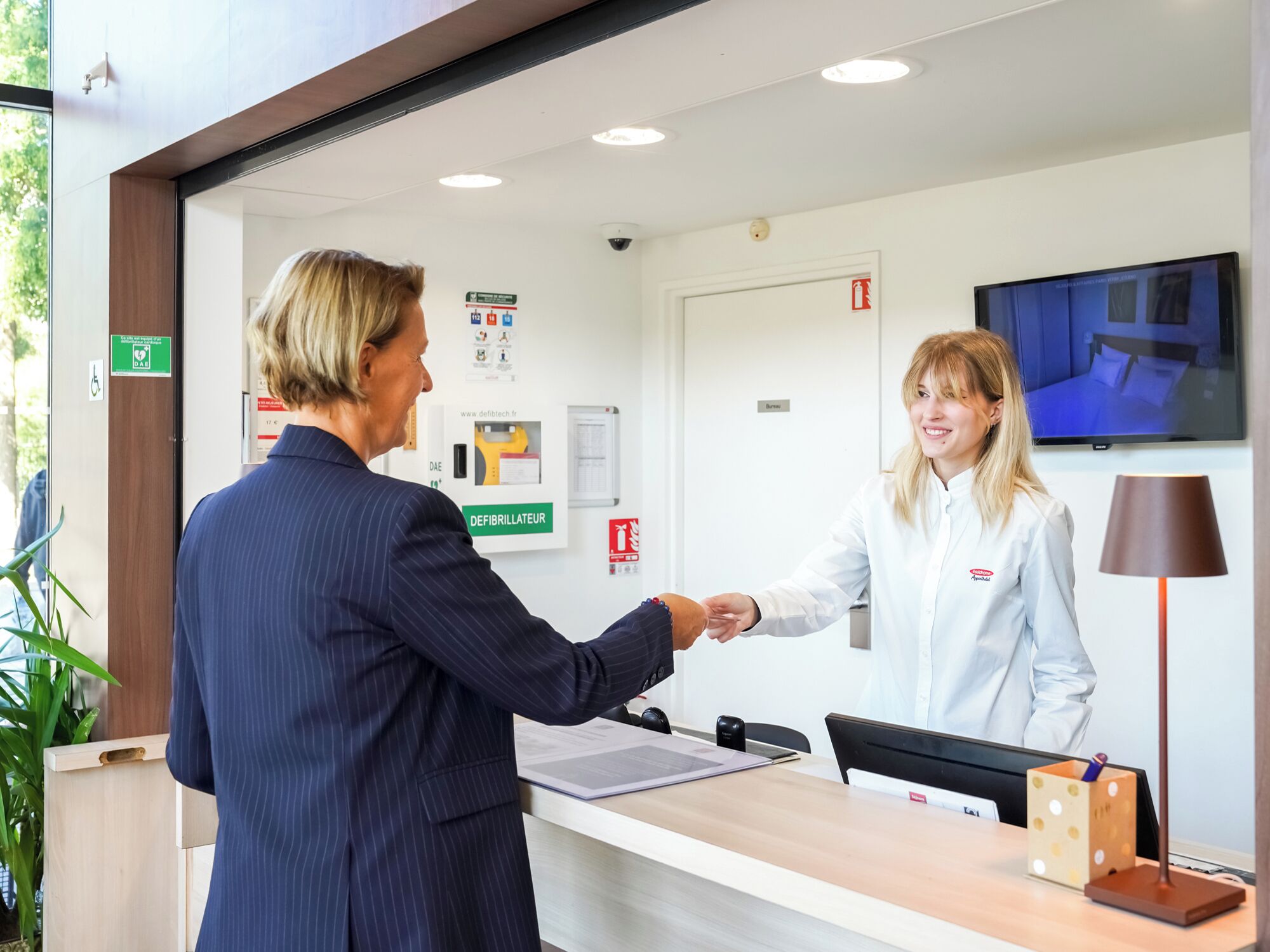 Una zona d'entrada amb taulell de recepció, un ordinador, detector d'incendis fixat a la paret i una pantalla de televisió.