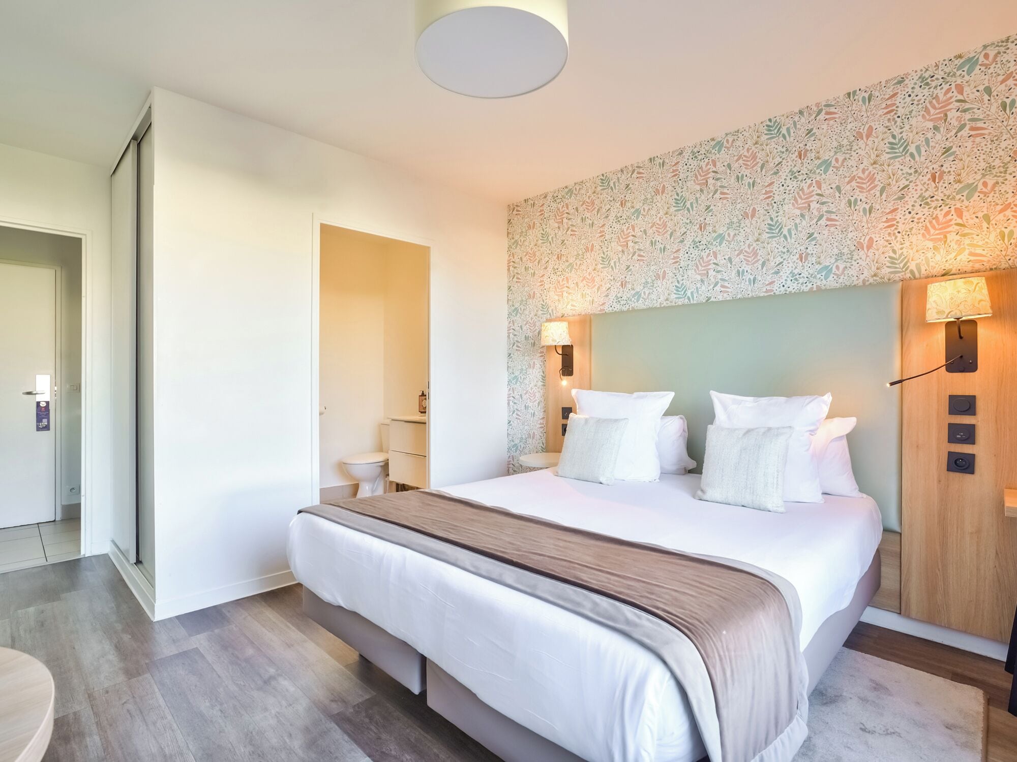 A modern hotel room with a large bed, a wooden headboard, and a floral-patterned wall.