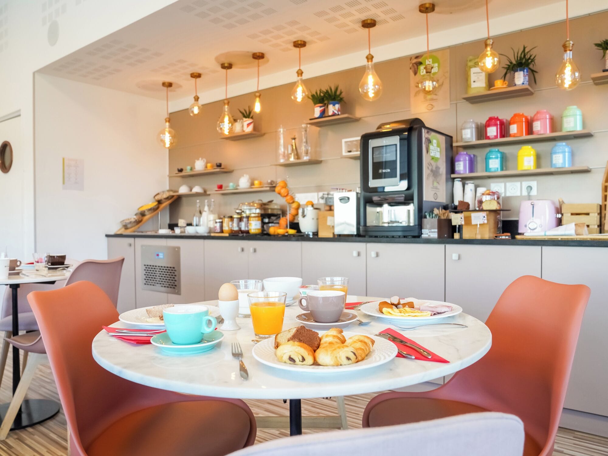 Una cuina moderna amb una màquina de cafè, prestatges amb recipients de colors i una zona de dinar amb taula rodona i cadires.