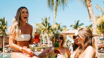 A poolside scene with two women enjoying drinks, surrounded by palm trees and umbrellas.