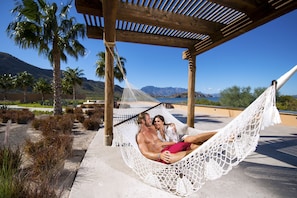 A hammock under a wooden pergola with a view of mountains and palm trees.