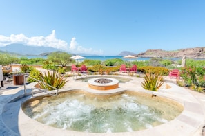 A hot tub surrounded by red chairs and a pool, with a view of the sea and mountains in the background.