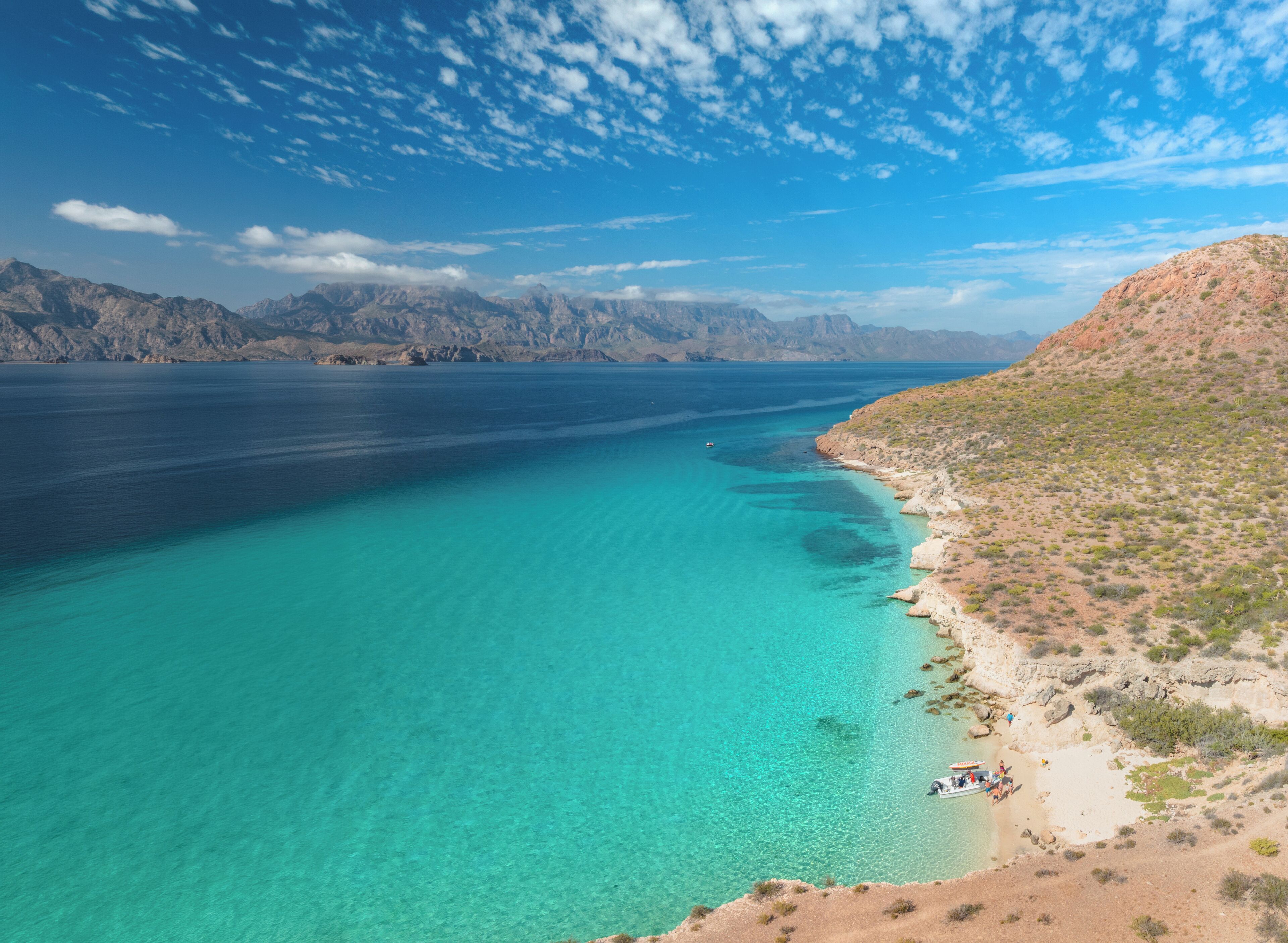 A coastal landscape with clear turquoise waters, a sandy beach, and rocky hills in the background.