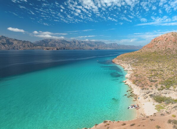 A coastal landscape with clear turquoise waters, a sandy beach, and rocky hills in the background.